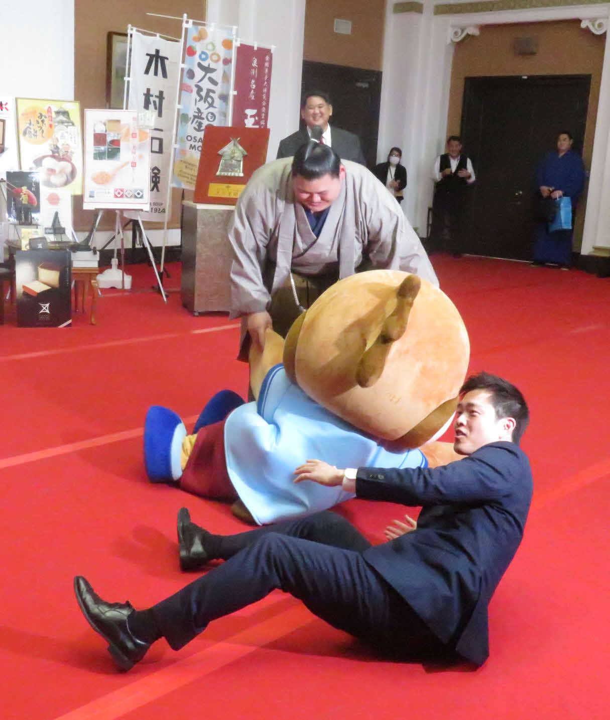 大の里（右）は要請を受けて相撲を取った、大阪府の公式キャラクター、もずやん（中央）を、加勢してきた大阪府の吉村知事（手前）とともに上手投げで仕留めた