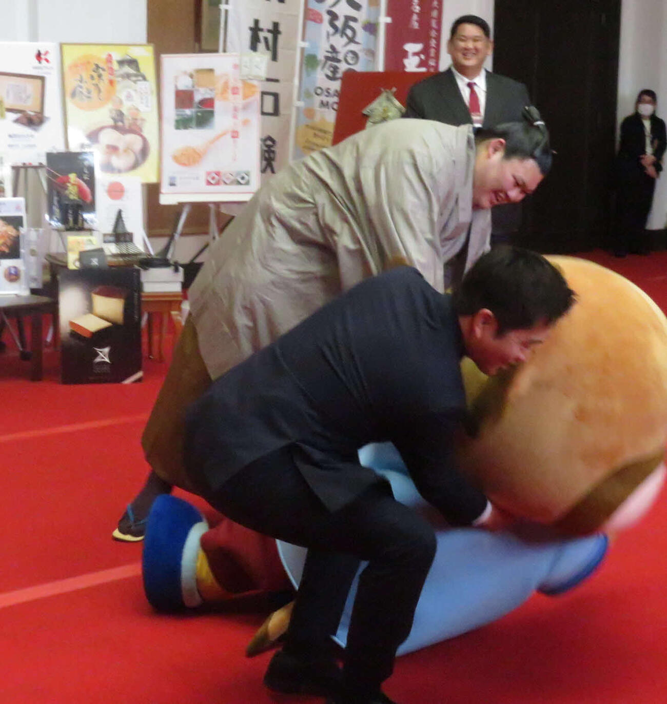 大の里（右）は要請を受けて相撲を取った、大阪府の公式キャラクター、もずやん（中央）を、加勢してきた大阪府の吉村洋文知事とともに上手投げで仕留めた。後方左は阿武松親方
