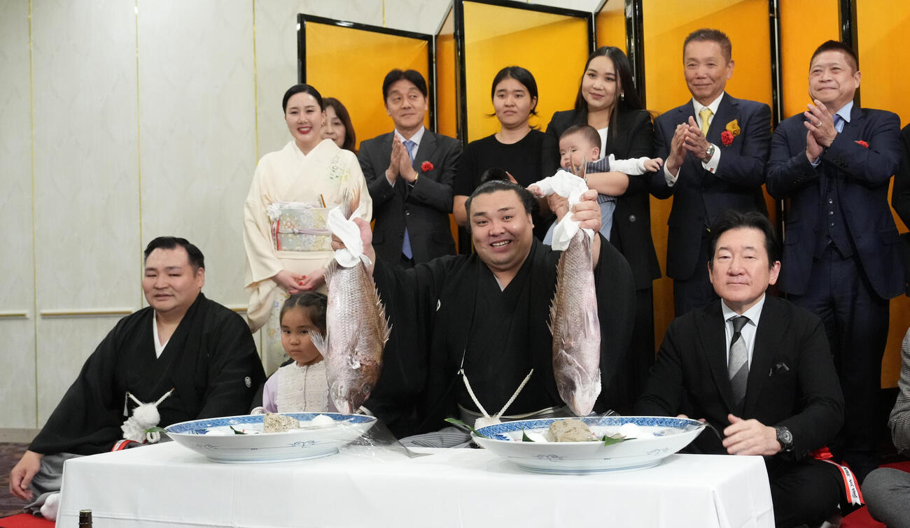 千秋楽パーティー会場で、左から師匠の音羽山親方、長女アヤゴーちゃん、後援会関係者らの祝福を受けながらお祝いのタイを持ち上げる霧島（中央）。後方左は音羽山夫人、霧島の後方右は長男トゥグドゥルくんを抱くホラン夫人＝2026年3月22日