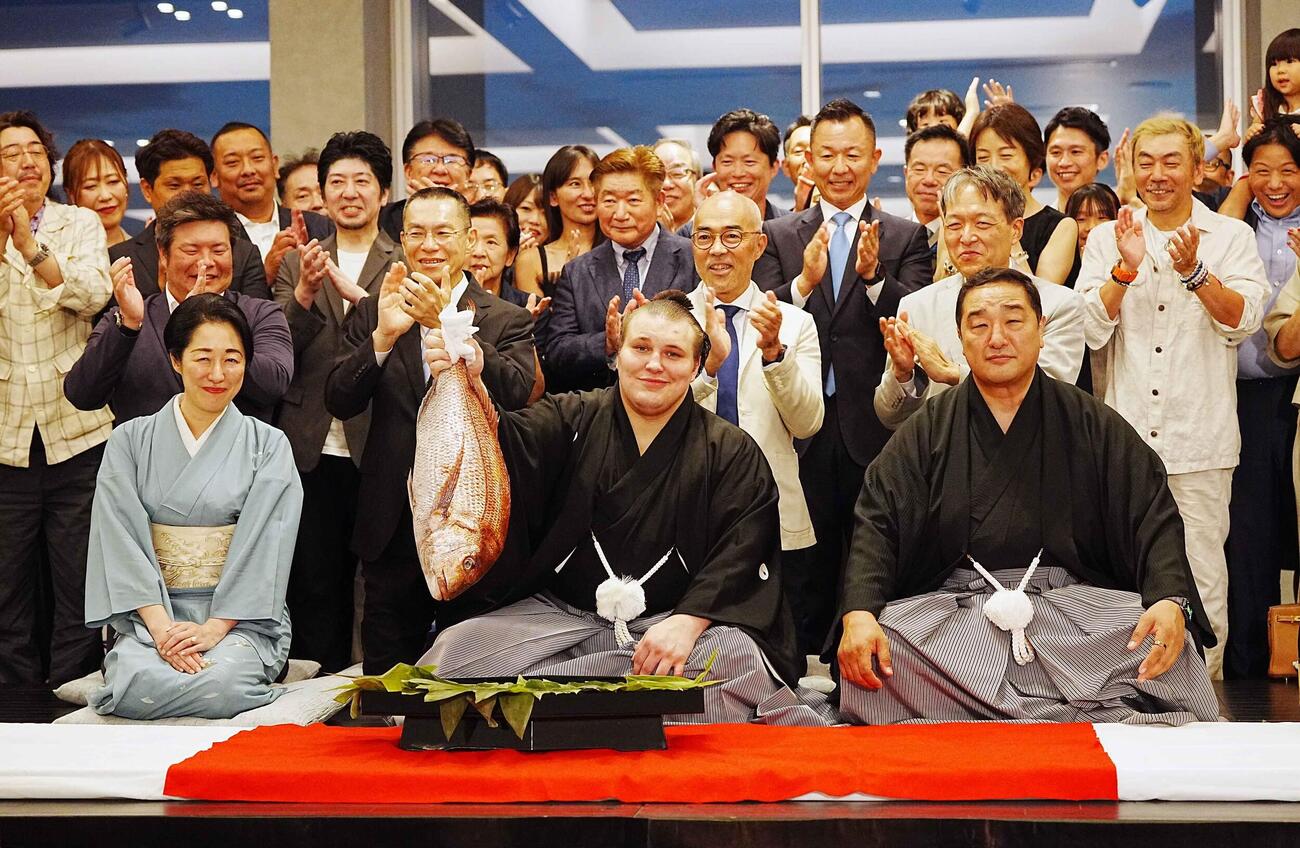 関係者の祝福を受けながらお祝いのタイを掲げる安青錦。右は師匠の安治川親方、左はおかみの絵莉さん（撮影・小沢裕）