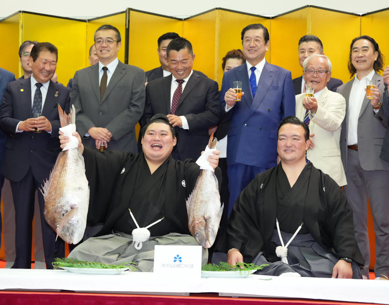 千秋楽パーティーで関係者の祝福を受けながらお祝いのタイを両手で持ち上げる大の里。右は師匠の二所ノ関親方（撮影・小沢裕）