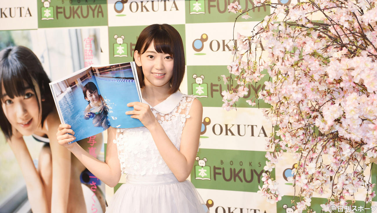 初写真集「さくら」の発売記念イベントを行ったHKT48の宮脇咲良は、お気に入りのページを開きながら桜の横で笑顔を見せる（2015年7月撮影）