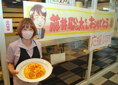 藤井聡太七段の似顔絵ポスターの横で特大オムライスを持つ「喫茶スマイル」の店主、鈴木松子さん(撮影・松浦隆司)