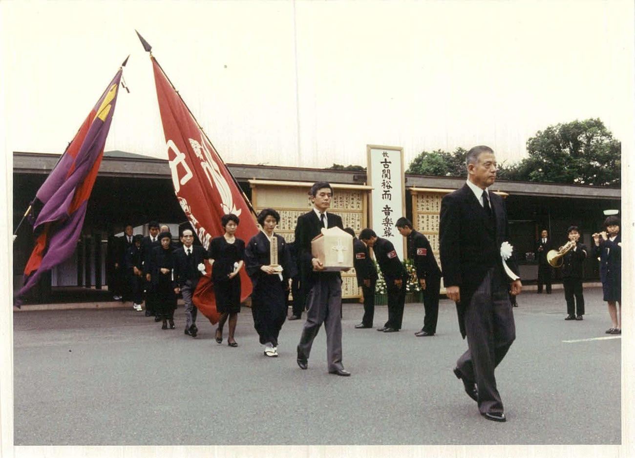 89年9月、青山葬儀場で行われた古関氏の音楽葬で、早大の校旗と慶大の三色旗が掲げられた。遺骨を持つのは長男正裕氏