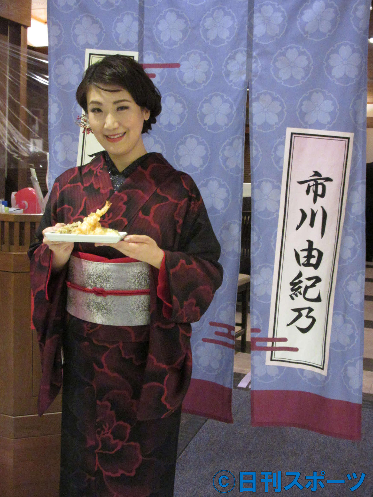 特注ののれんの前に天ぷらを持って立つ市川由紀乃（東京・新宿つな八京王店）