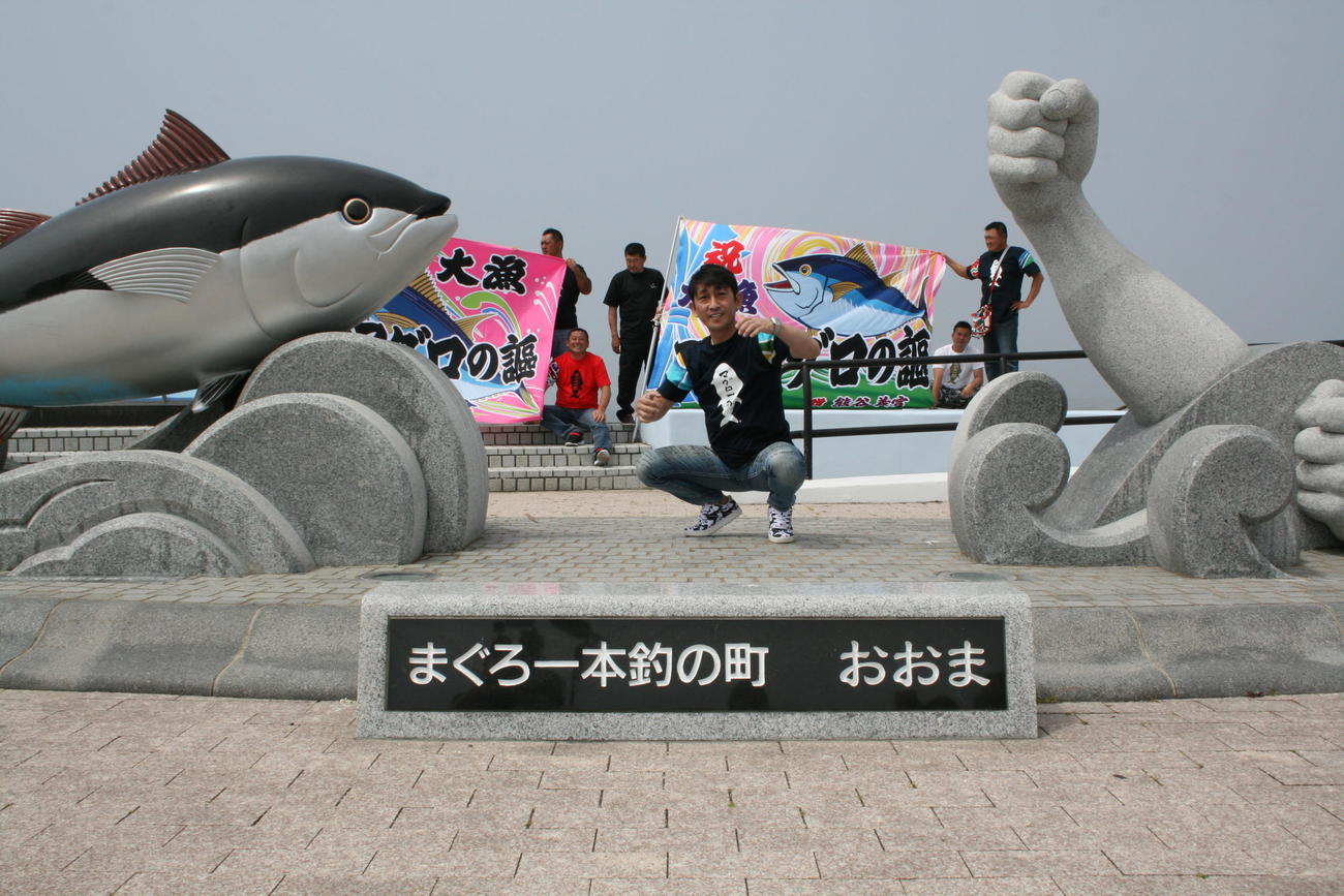 マグロのモニュメントと大漁旗をバックにポーズを取る福田こうへい