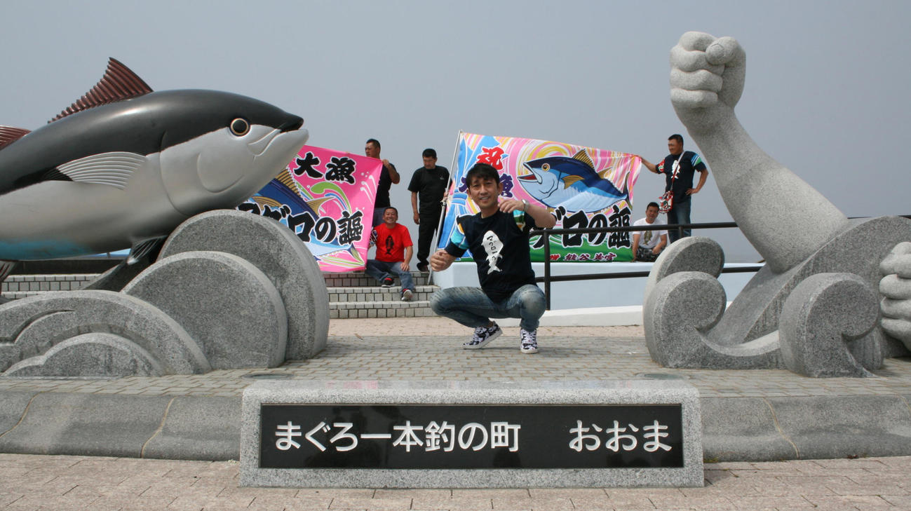 マグロのモニュメントと大漁旗をバックにポーズを取る福田こうへい