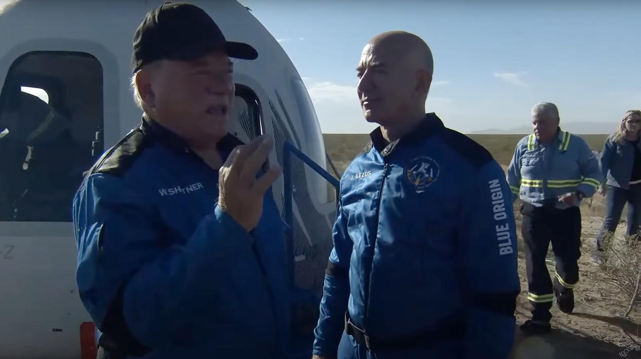 宇宙空間に到達後、地上に帰還し、ジェフ・ベゾス氏（中央）と話すウィリアム・シャトナーさん（左）。奥は宇宙船ニューシェパード（ブルーオリジンの中継動画から＝共同）