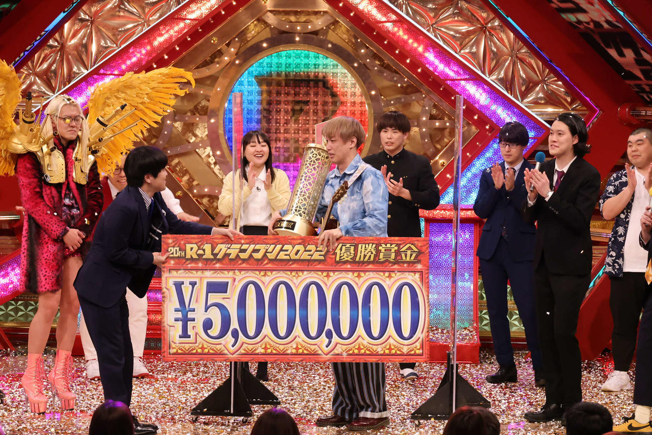 「R－1グランプリ2022」を制したお見送り芸人しんいち（中央）はバカリズム（左手前）から優勝賞金の目録を受けとる。左後方は敗れたZAZY（C）カンテレ