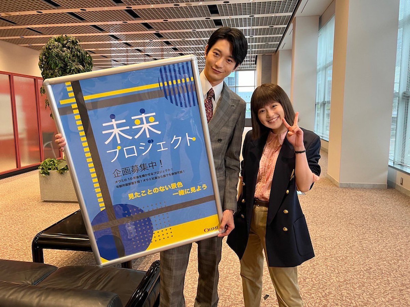 未来プロジェクトのパネルを持った向井理（左）と隣でピースサインをする今田美桜（「悪女（わる）」オフィシャルブログより）