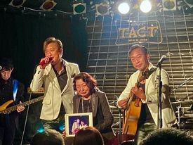 ジョニーさんと自身のツーショットの写真を持ったマリーさん（中）を囲んで「二人だけ」を歌うケニー（左）と弘也