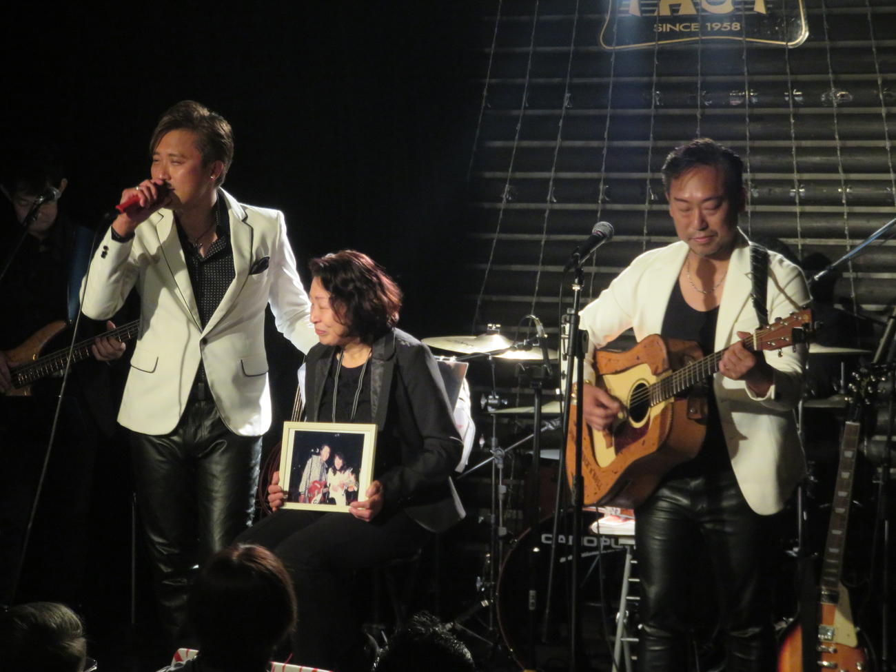 ジョニーさんと自身のツーショットの写真を持ったマリーさん（中）を囲んで「二人だけ」を歌うケニー（左）と弘也