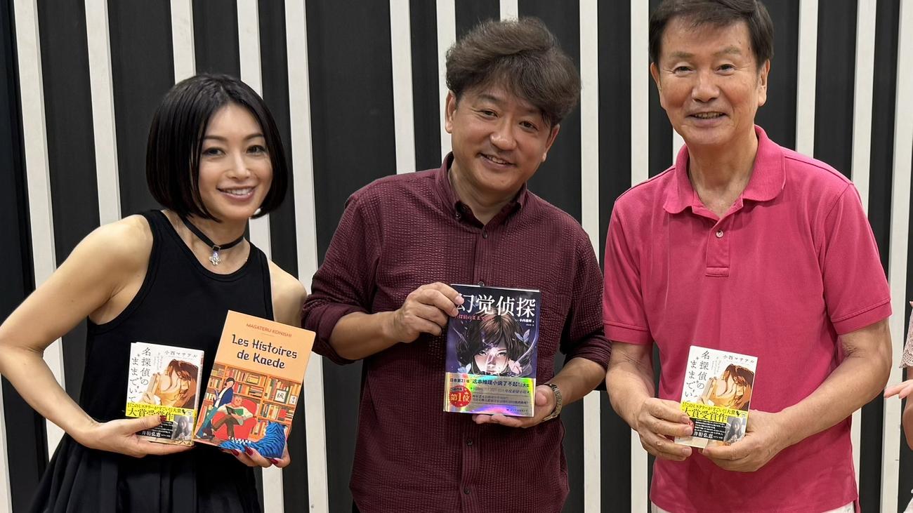 中国でも「幻覚探偵」と題して出版された「名探偵のままでいて」を持つ小西マサテル氏（中）。左は酒井法子、右は森田健作