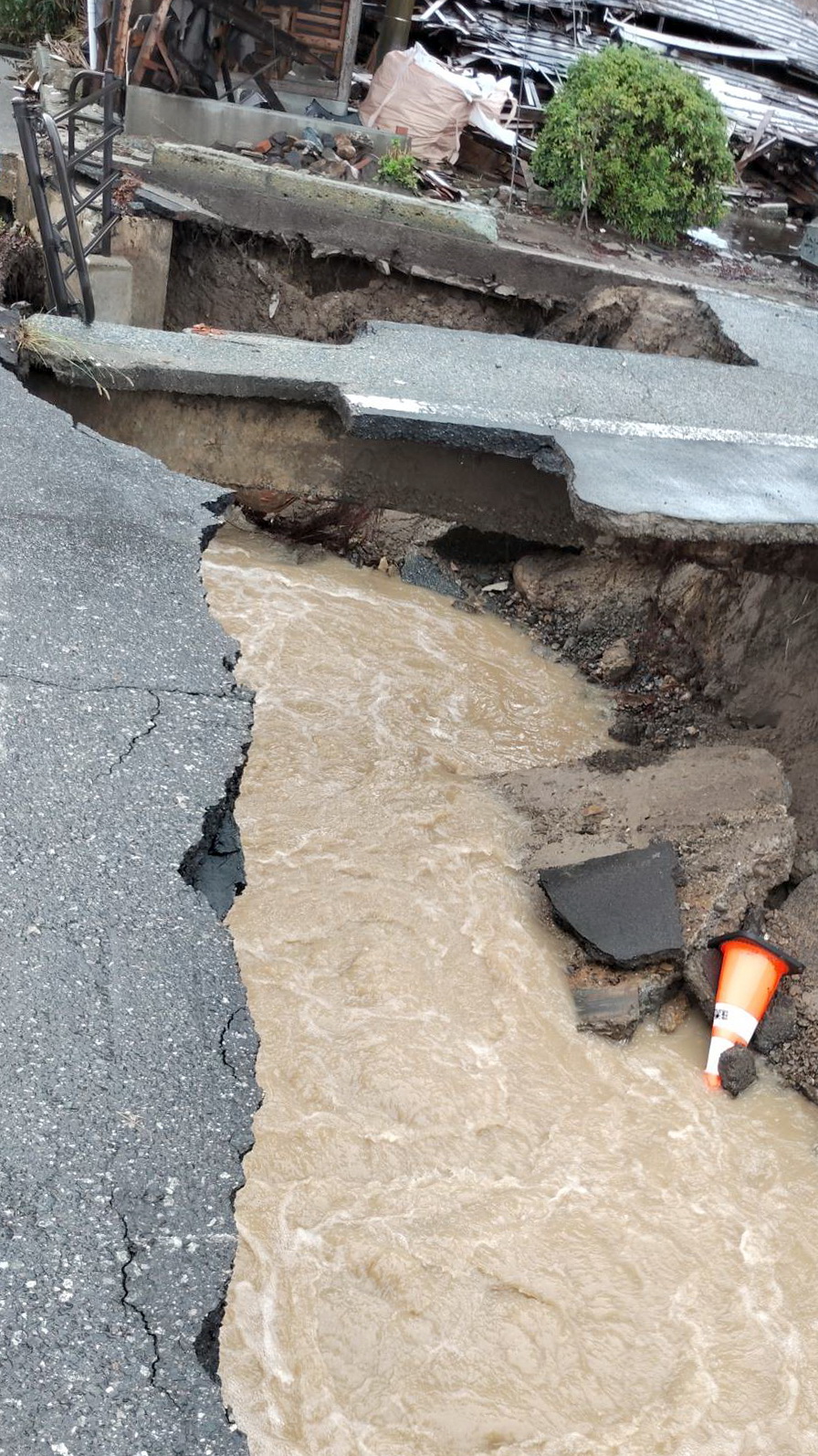大雨の影響で陥没した石川・珠洲市の道路や歩道に流れ込む濁流。奥には倒壊した家屋も（出村正幸さん提供）