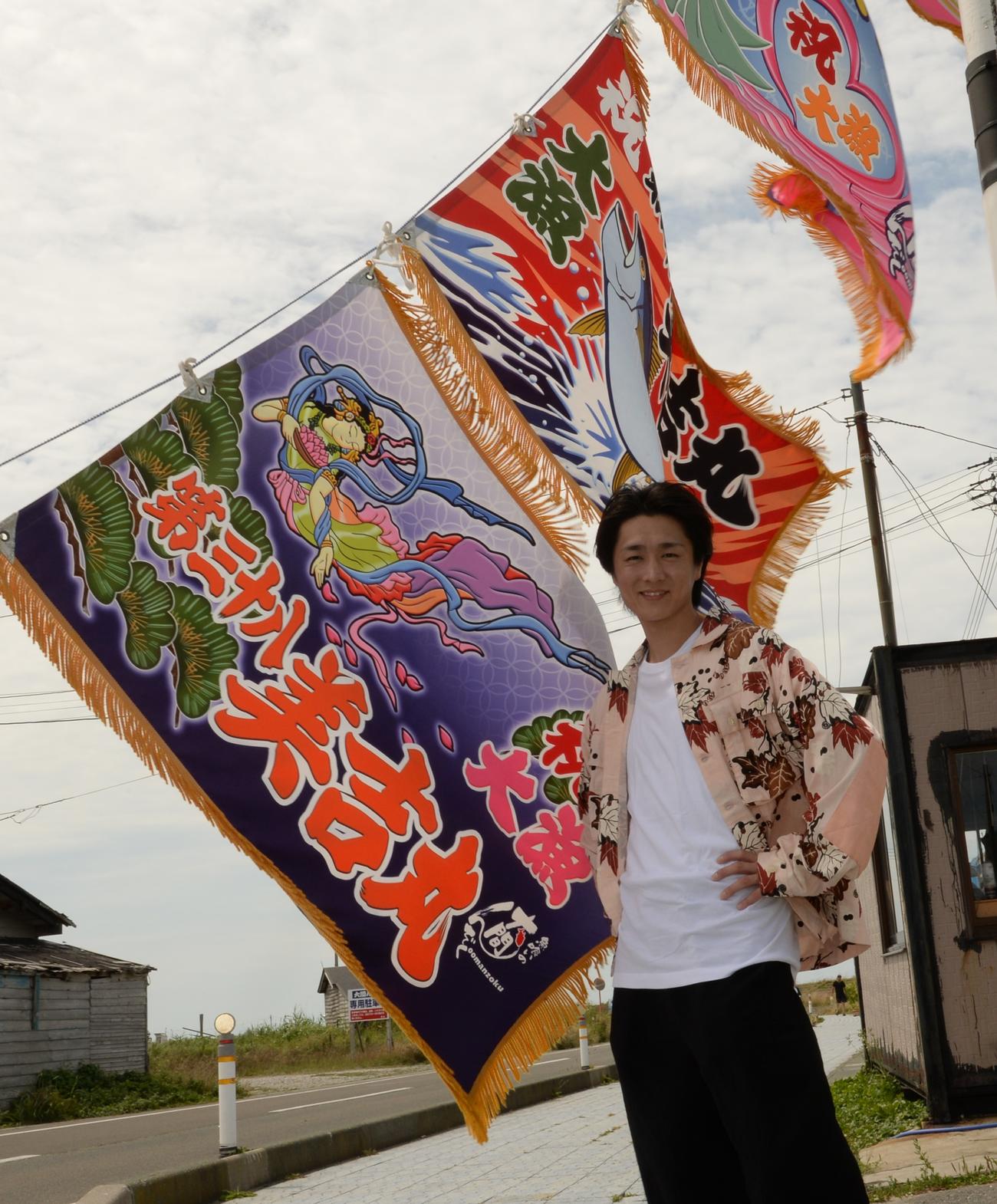 大漁旗をバックにポーズを決める真田ナオキ