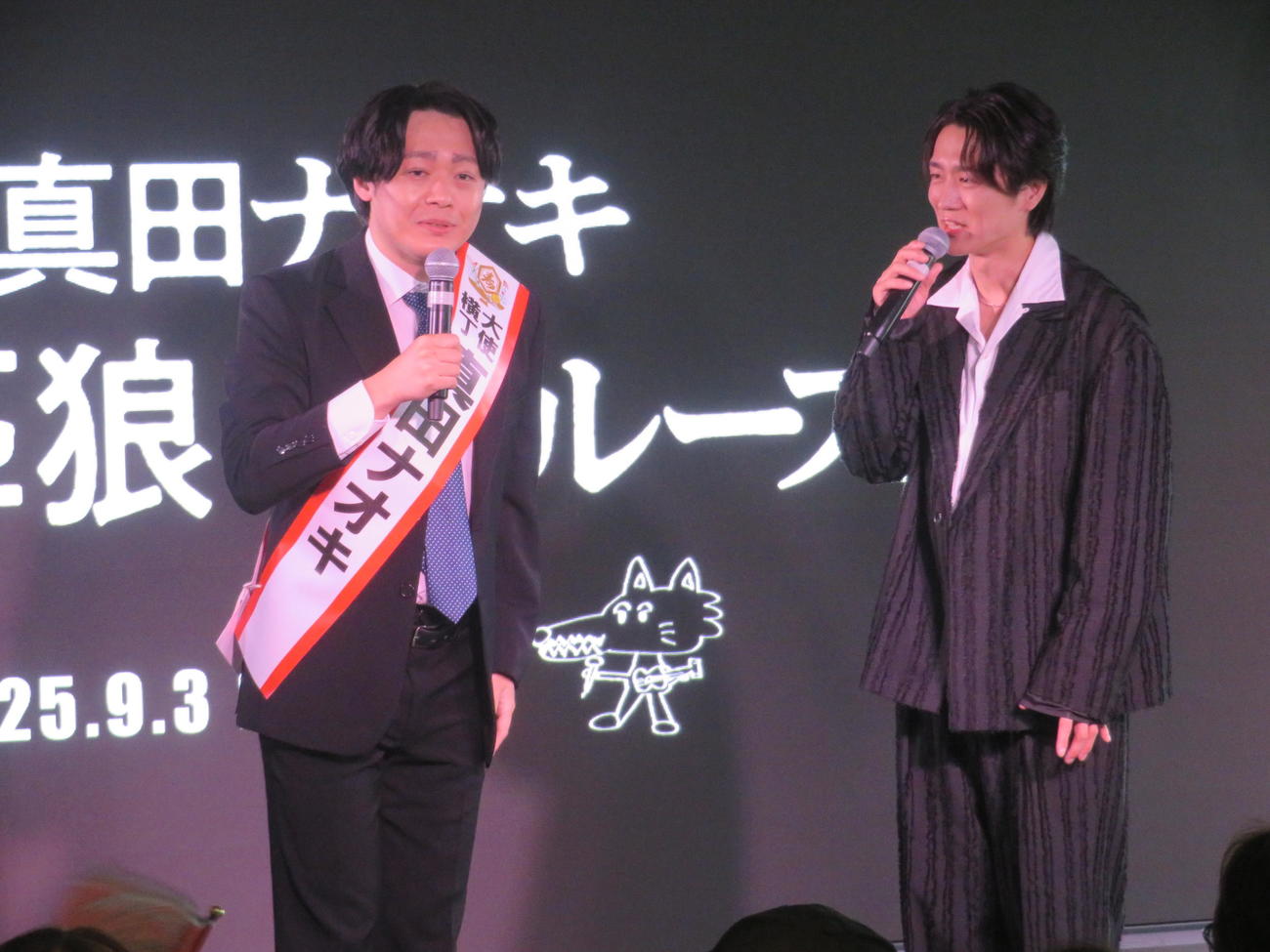 ものまね芸人レッツゴーよしまさ（左）にタスキをプレゼントした真田ナオキ（撮影・川田和博）