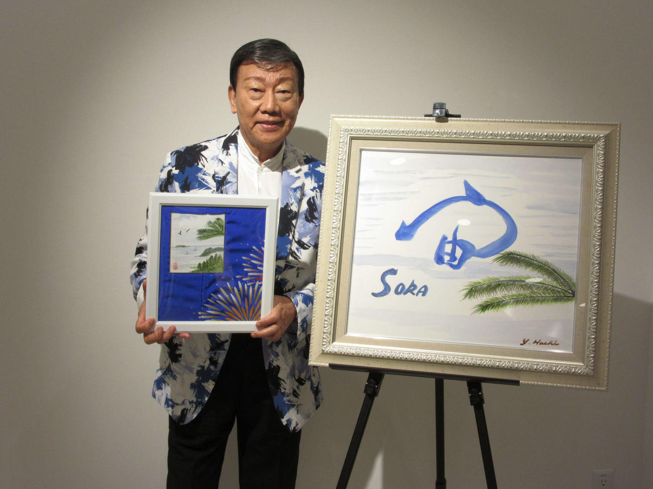 初の書画個展を開催した橋幸夫さん。持っているのが絵画「花火音頭」、右が書画展のテーマの書「宙（そら）」
