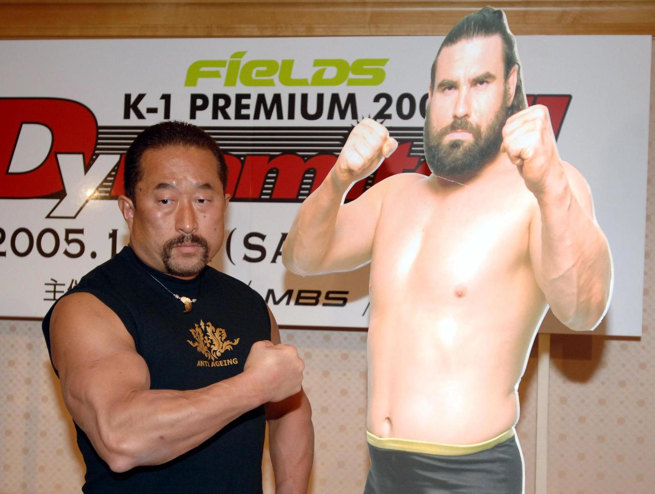 2005年12月、K−1 Dynamite記者会見　対戦相手に決まったザ・プレデターのパネルの横でポーズをとる角田信朗