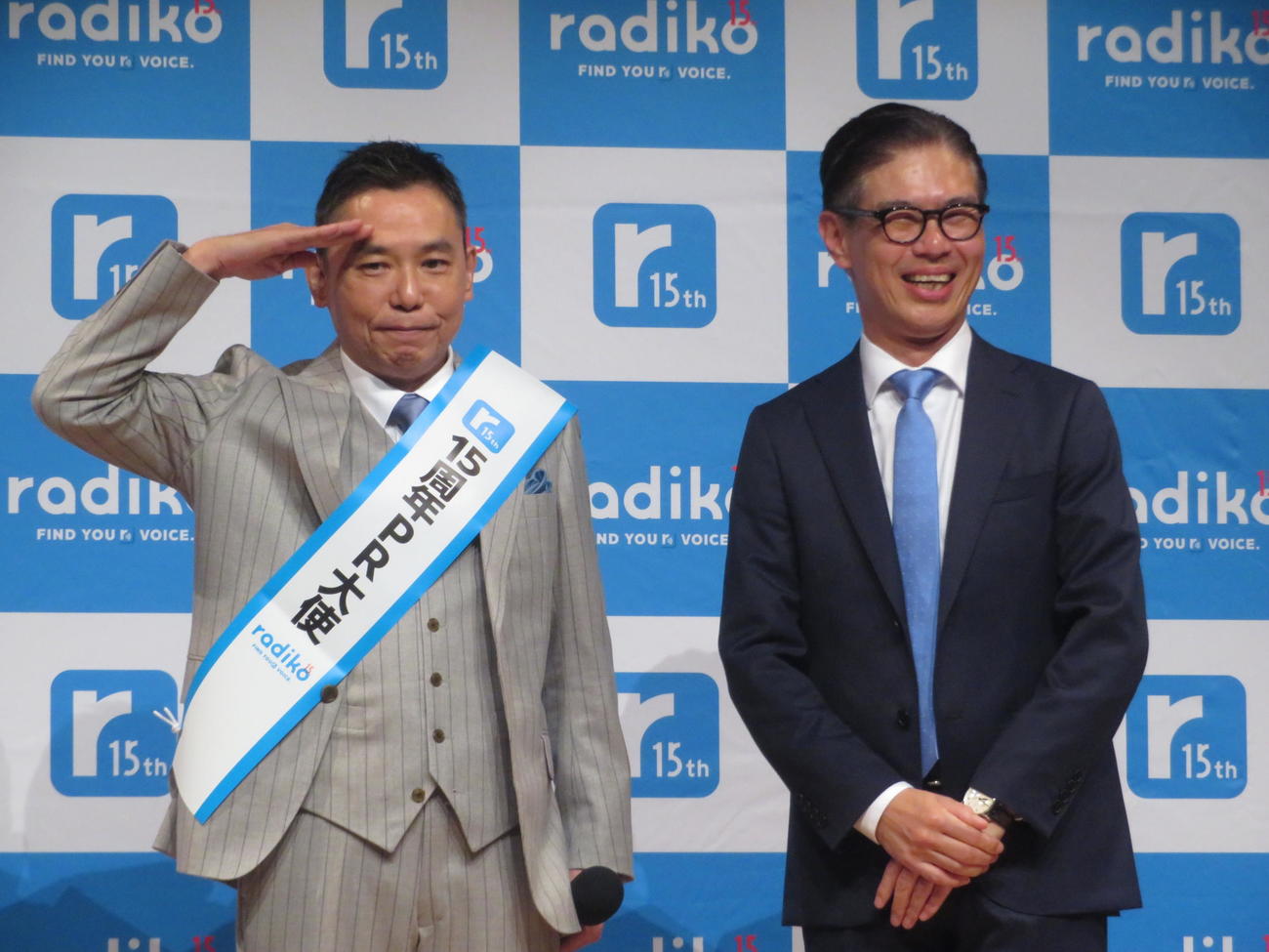 池田社長からradiko15周年PR大使のタスキをかけられ敬礼する太田光（左）（撮影・川田和博）
