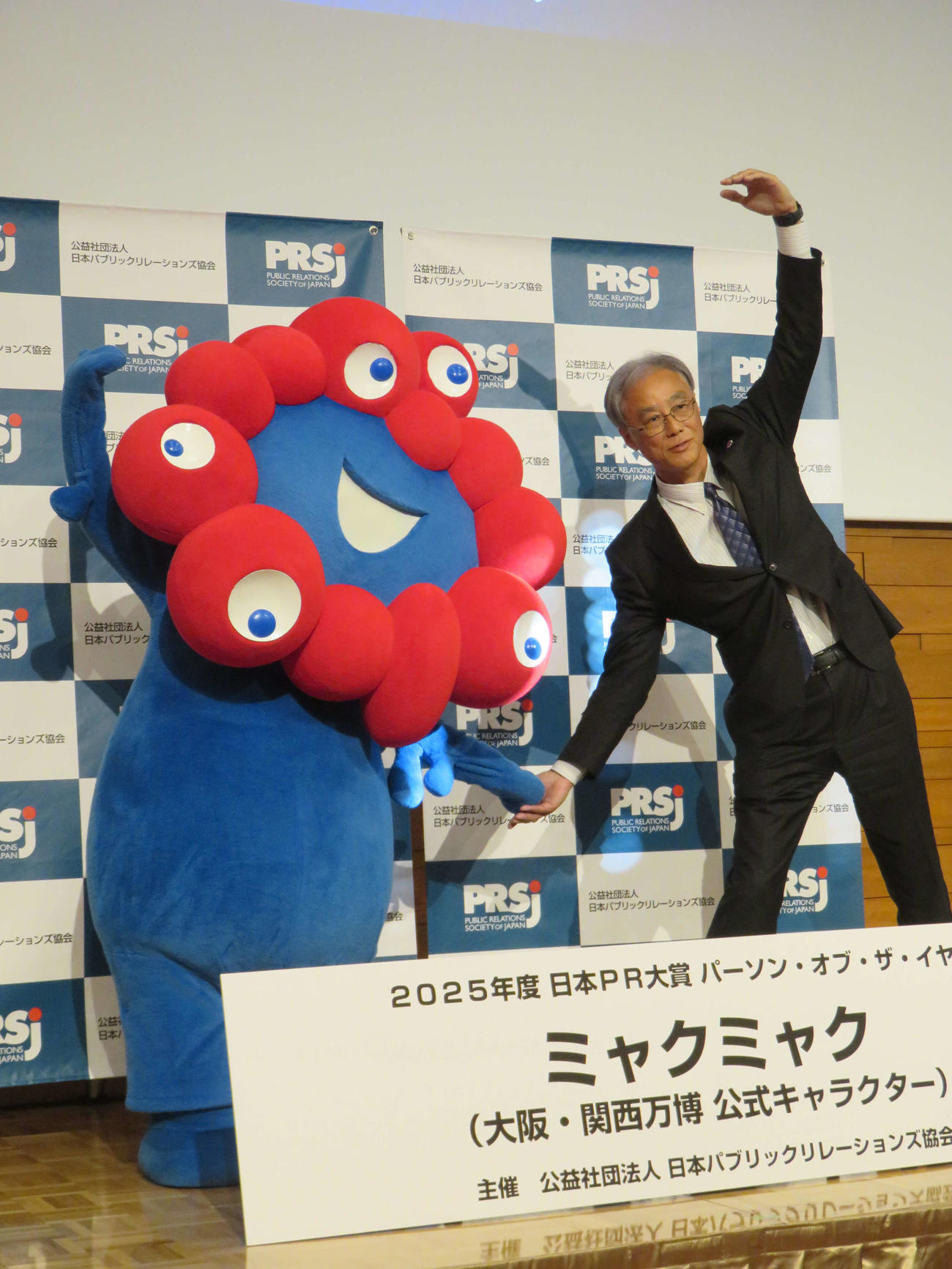 2025年度日本PR大賞でパーソン・オブ・ザ・イヤーを受賞し、いのちのポーズをするミャクミャク（左）と2025年日本国際博覧会協会の副事務総長・高科淳氏
