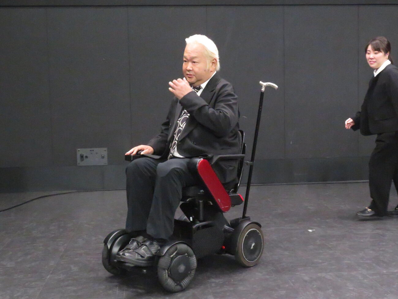 「死ぬまでにやりたいことプロジェクト」の概要を発表した会見を終え、電動車いすで引き揚げるハチミツ二郎（左）。右は吉本興業の山岸未歩さん