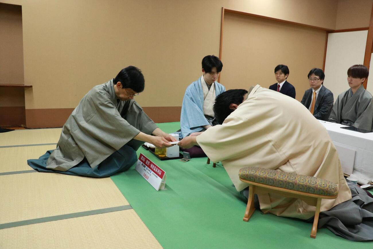 立会人の谷川浩司17世名人（左）に封じ手の封筒を渡す糸谷哲郎九段（右）。中央は藤井聡太名人（日本将棋連盟提供）