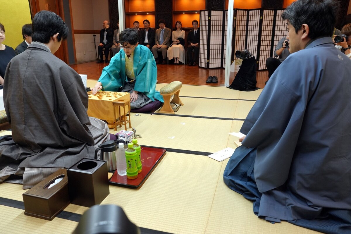 立会人の森内俊之九段（右）が伊藤匠2冠（中央）の封じ手を開封して2日目が再開された（左は藤井聡太王位、日本将棋連盟提供）