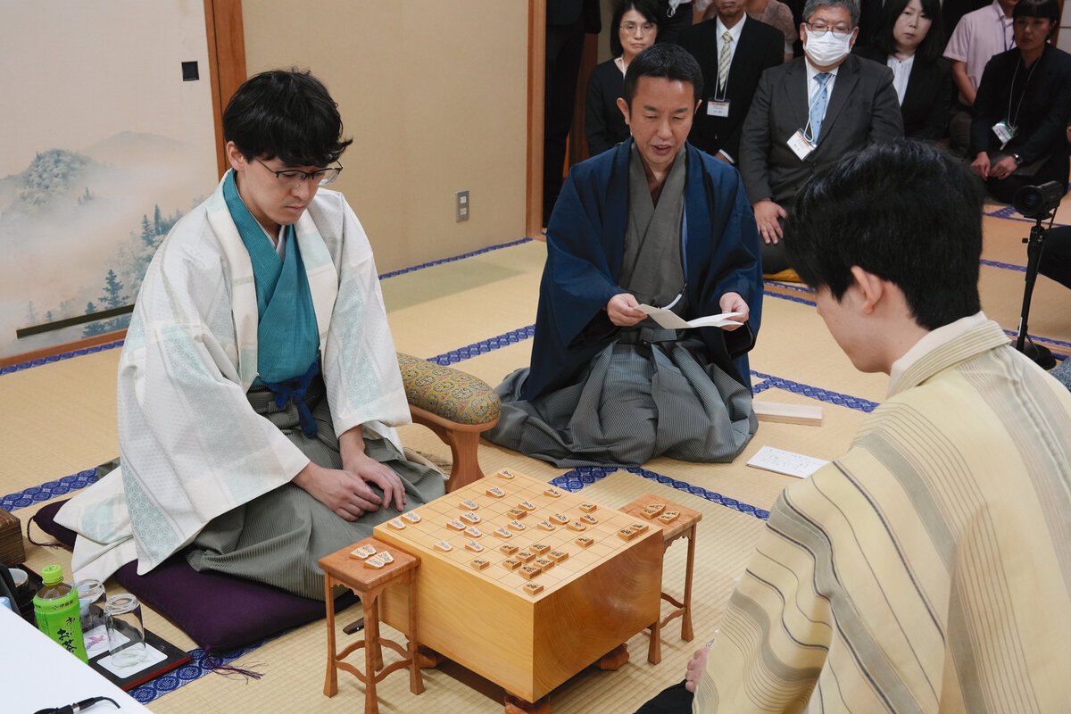 伊藤匠2冠（左）の封じ手を立会人の屋敷伸之九段（中央）が読み上げて王位戦第3局は2日目がスタート（右は藤井聡太王位、日本将棋連盟提供）