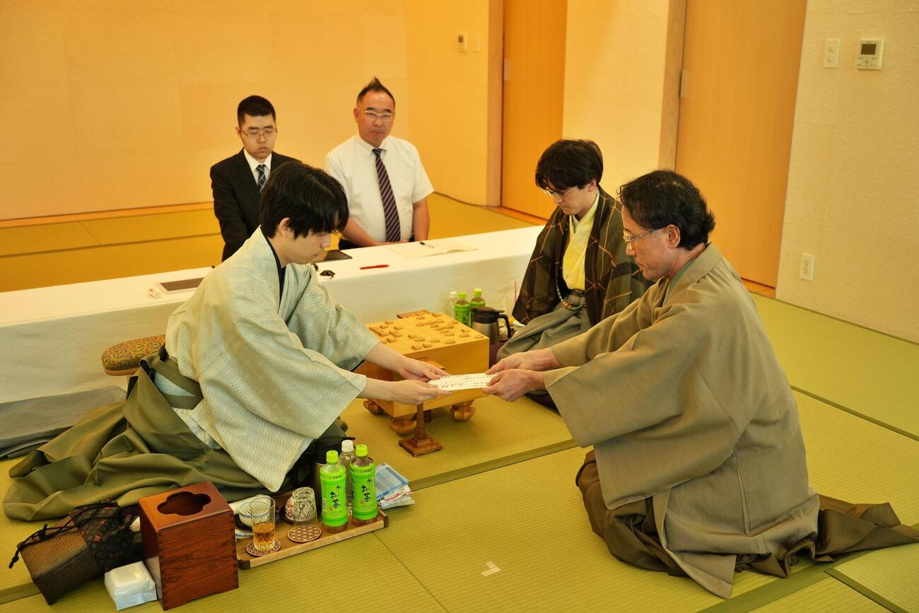封じ手の封筒の立会人の深浦康市九段（右）に手渡す藤井聡太王位（左）（日本将棋連盟提供）