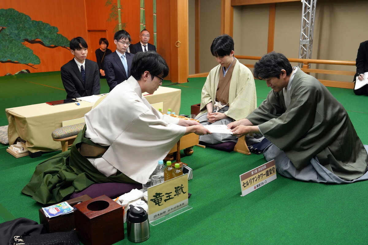 服部慎一郎七段（左）が立会人の森内俊之九段（右）に封じ手の入った封筒を渡して竜王戦第1局の初日は終了（中央は藤井聡太竜王、日本将棋連盟提供）