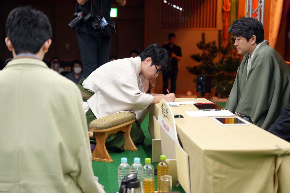 藤井聡太竜王（左）が見つめる前で服部慎一郎七段（中央）は立会人の森内俊之九段（右）に教えてもらいながら封じ手を行う（日本将棋連盟提供）
