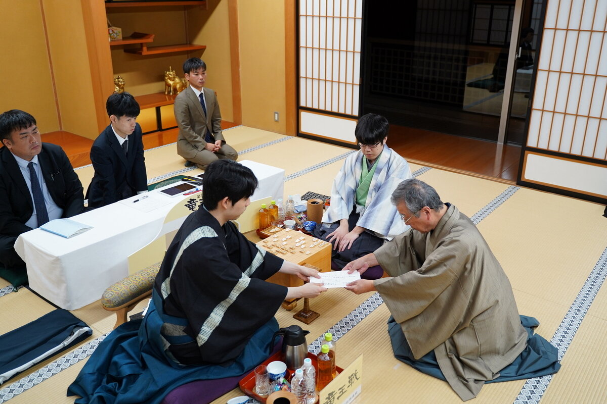 藤井聡太竜王（左）は立会人の青野照市九段（右）に封じ手の入った封筒を渡す（中央は服部慎一郎七段、日本将棋連盟提供）