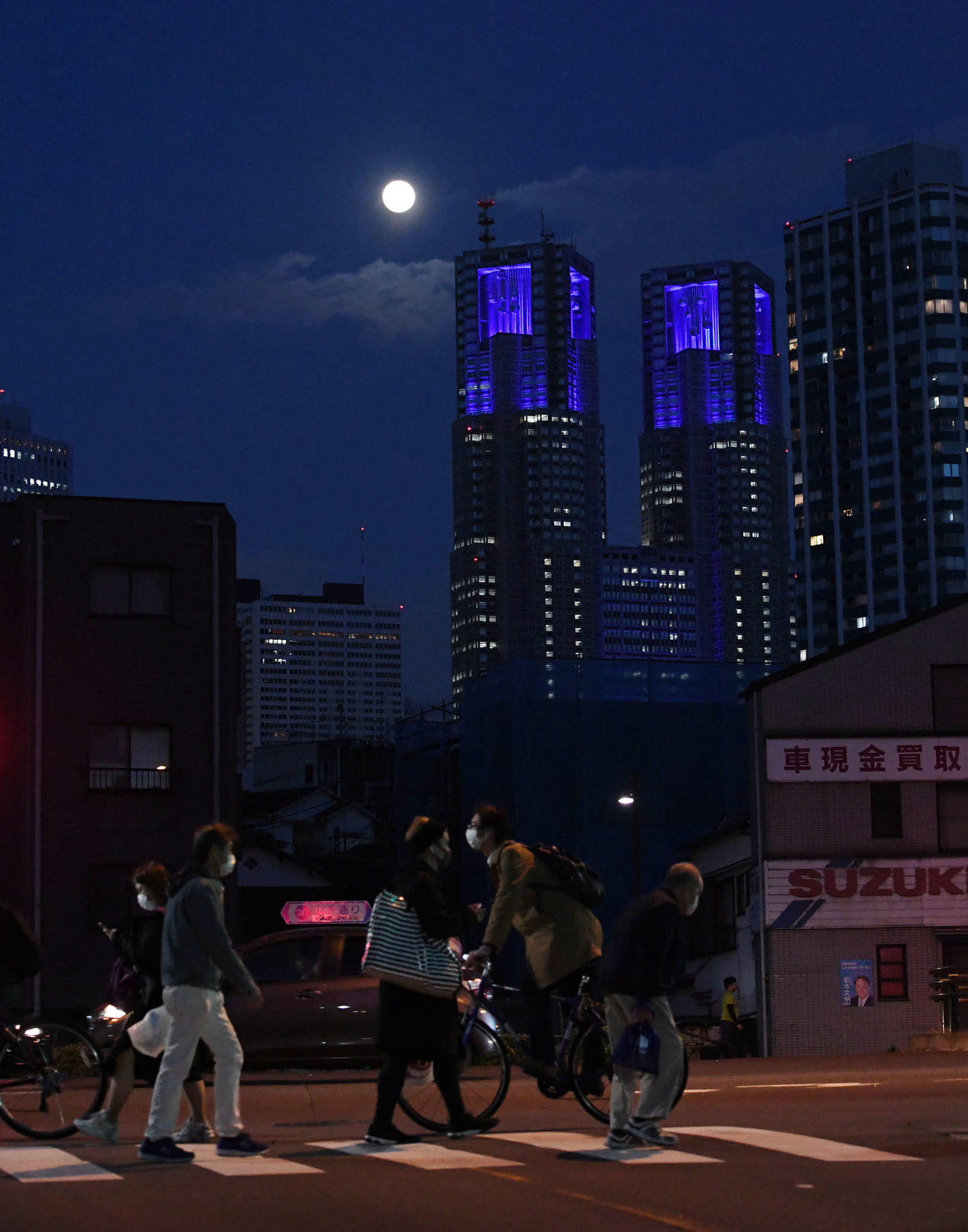 月に照らされる東京都庁（後方中央）。手前はマスク着用で帰宅を急ぐ人たち（撮影・山崎安昭）