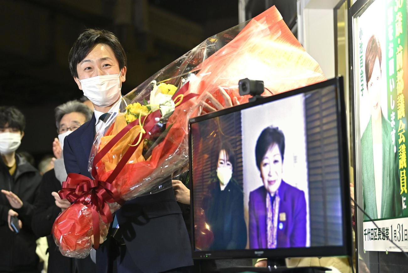 東京都千代田区長選で当選確実となり、花束を受け取った樋口高顕氏。モニター画面右はオンラインで祝福する小池百合子知事（共同）