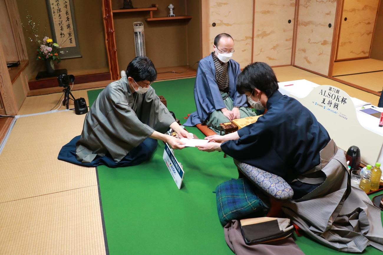 立会人の谷川浩司九段（左）に封じ手の封筒を手渡す藤井聡太竜王（日本将棋連盟提供）
