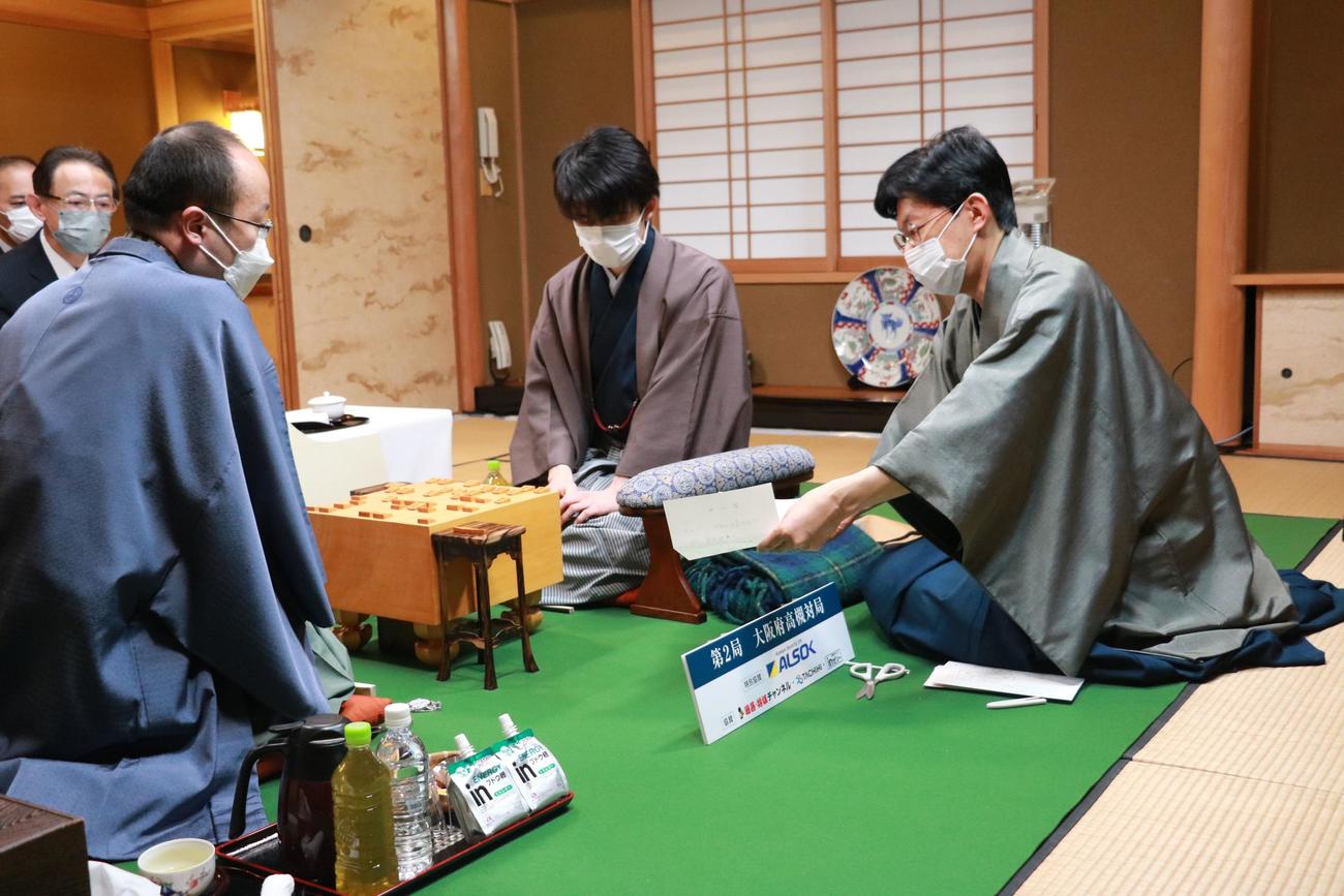 立会人の谷川浩司九段が示す封じ手を確認する藤井聡太竜王（右）、渡辺明王将（日本将棋連盟提供）