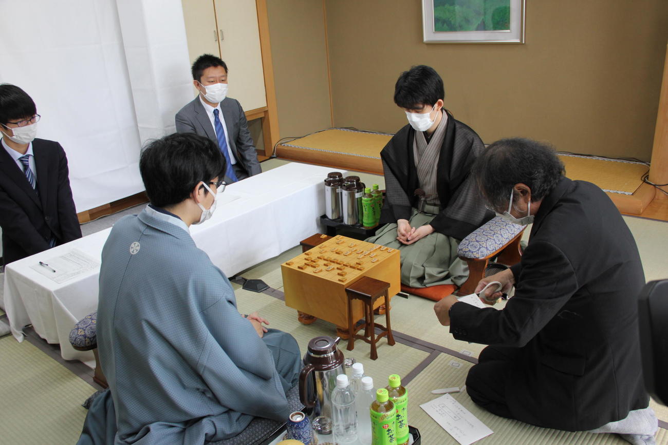 立会人の福崎文吾九段が封じ手の封筒を開封するのを待つ藤井聡太王位（右）豊島将之九段（日本将棋連盟提供）
