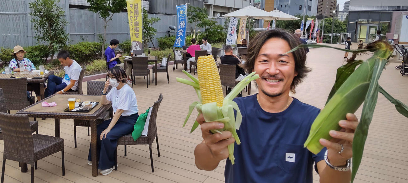 築地魚河岸3階のテラス席で自ら収穫したトウモトコシを手に笑顔の石川直宏さん（撮影・寺沢卓）