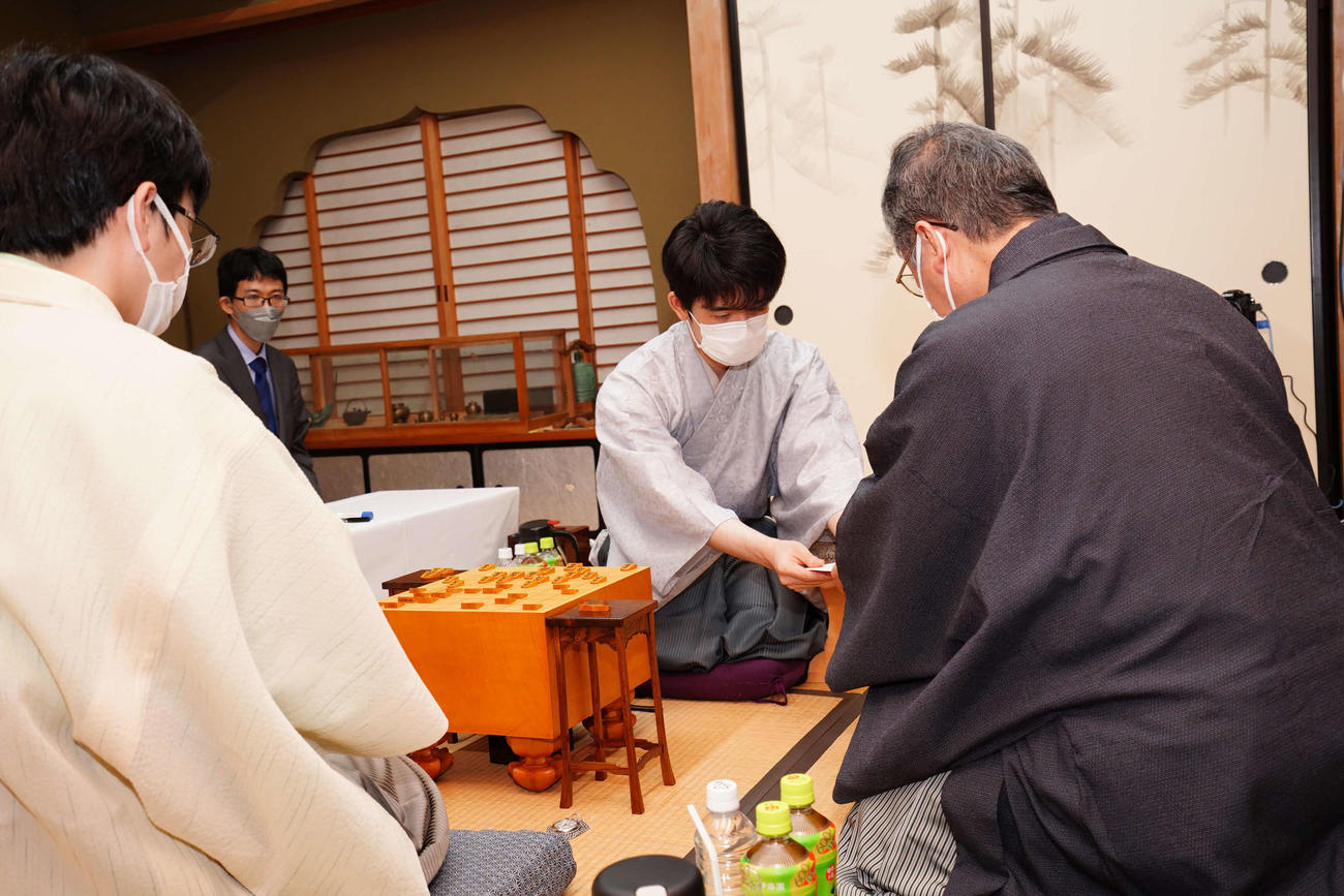 封じ手の入った封筒を立会人の青野照市九段（右）に手渡す藤井聡太王位（中央、左は豊島将之九段、日本将棋連盟提供）