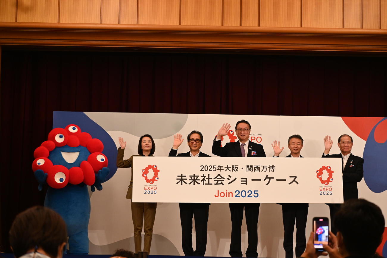 2025年の大阪・関西万博の公式キャラクター「ミャクミャク」（左）は写真撮影で岡田直樹・国際博覧会担当大臣（右から3人目）と代表者らをエスコート（撮影・松浦隆司）