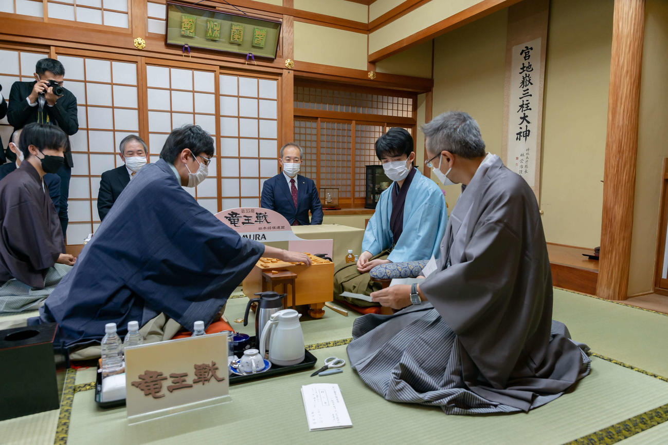 立会人の中村修九段（手前）が広瀬章人八段（左）の封じ手を示し、確認する藤井聡太竜王（右）（日本将棋連盟提供）