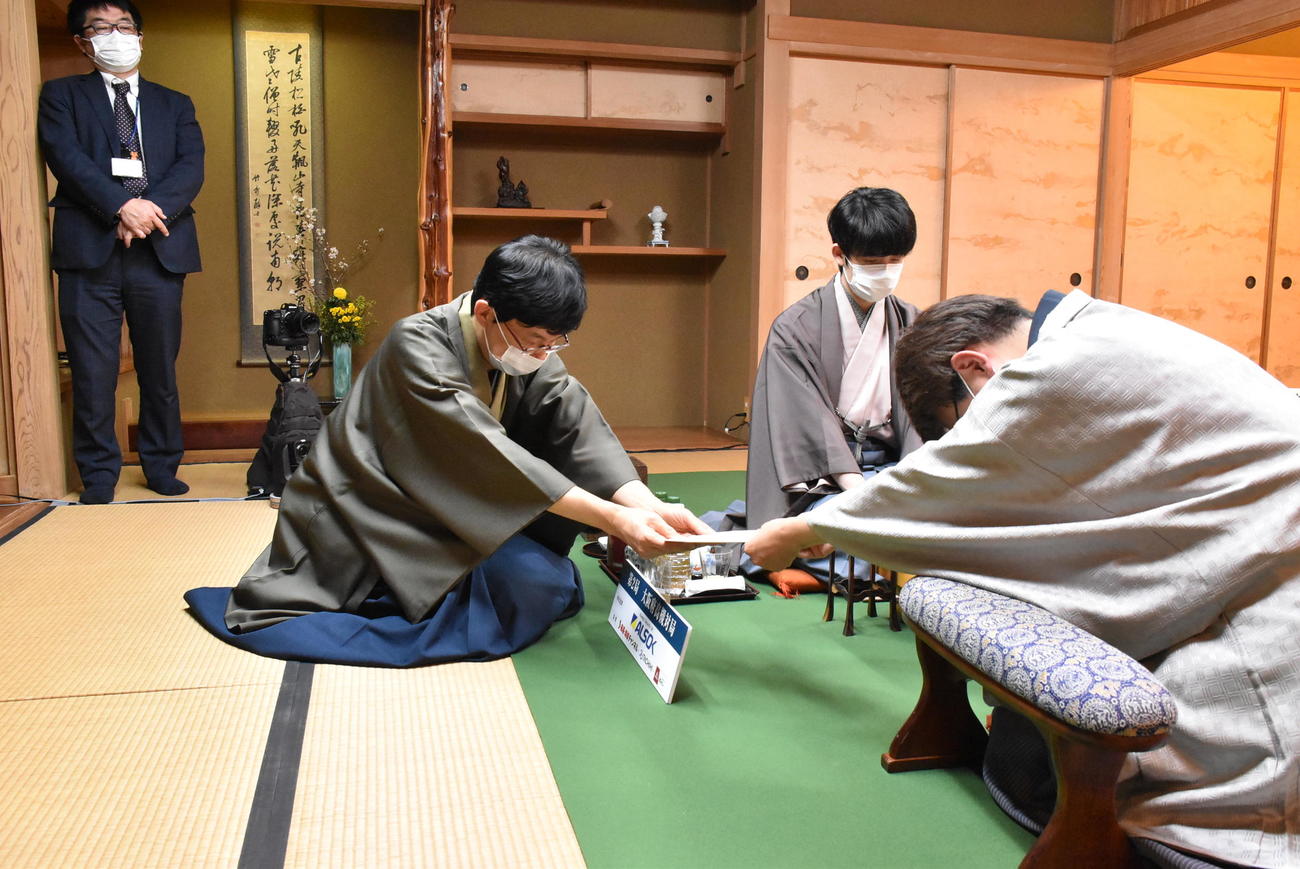 立会人の谷川浩司17世名人（左）に封じ手の封筒を手渡す羽生善治九段（右）、奥は藤井聡太王将（日本将棋連盟提供）