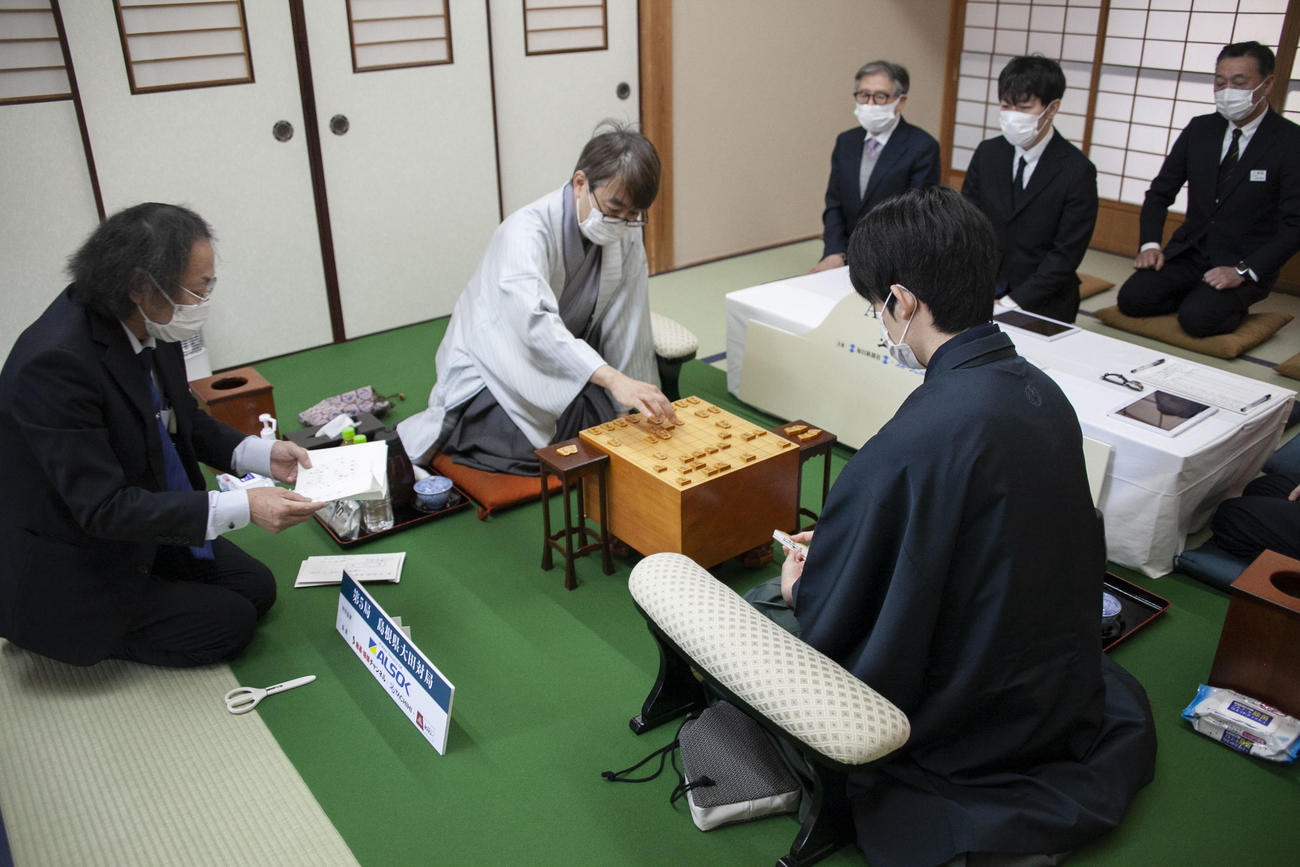 立会人の福崎文吾九段（左）が封じ手を読み上げ、封じ手を指す羽生善治九段（中央）、手前は藤井聡太王将（日本将棋連盟提供）