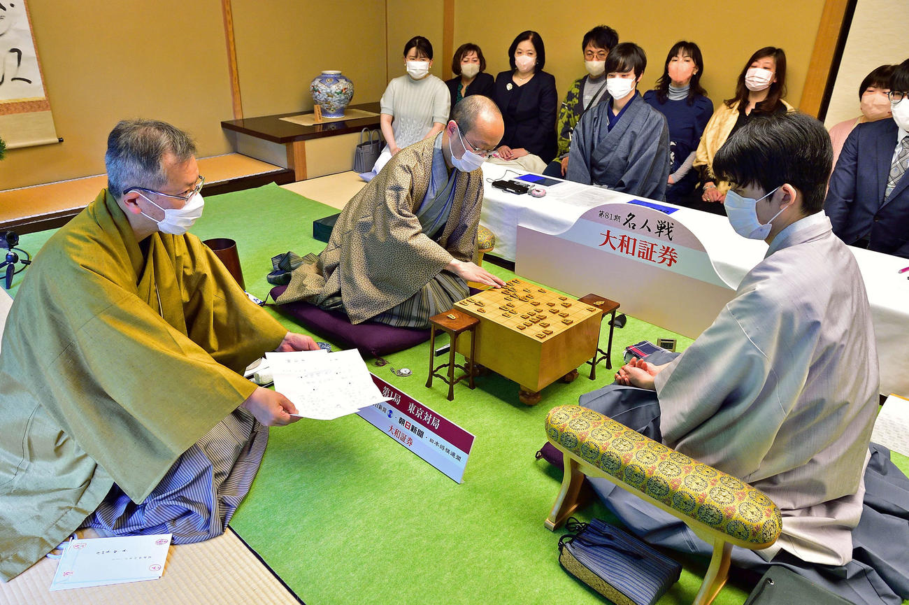 立会人の中村修九段（左）が渡辺明名人（中央）の封じ手を開封して名人戦第1局は再開（右は藤井聡太竜王、日本将棋連盟提供）