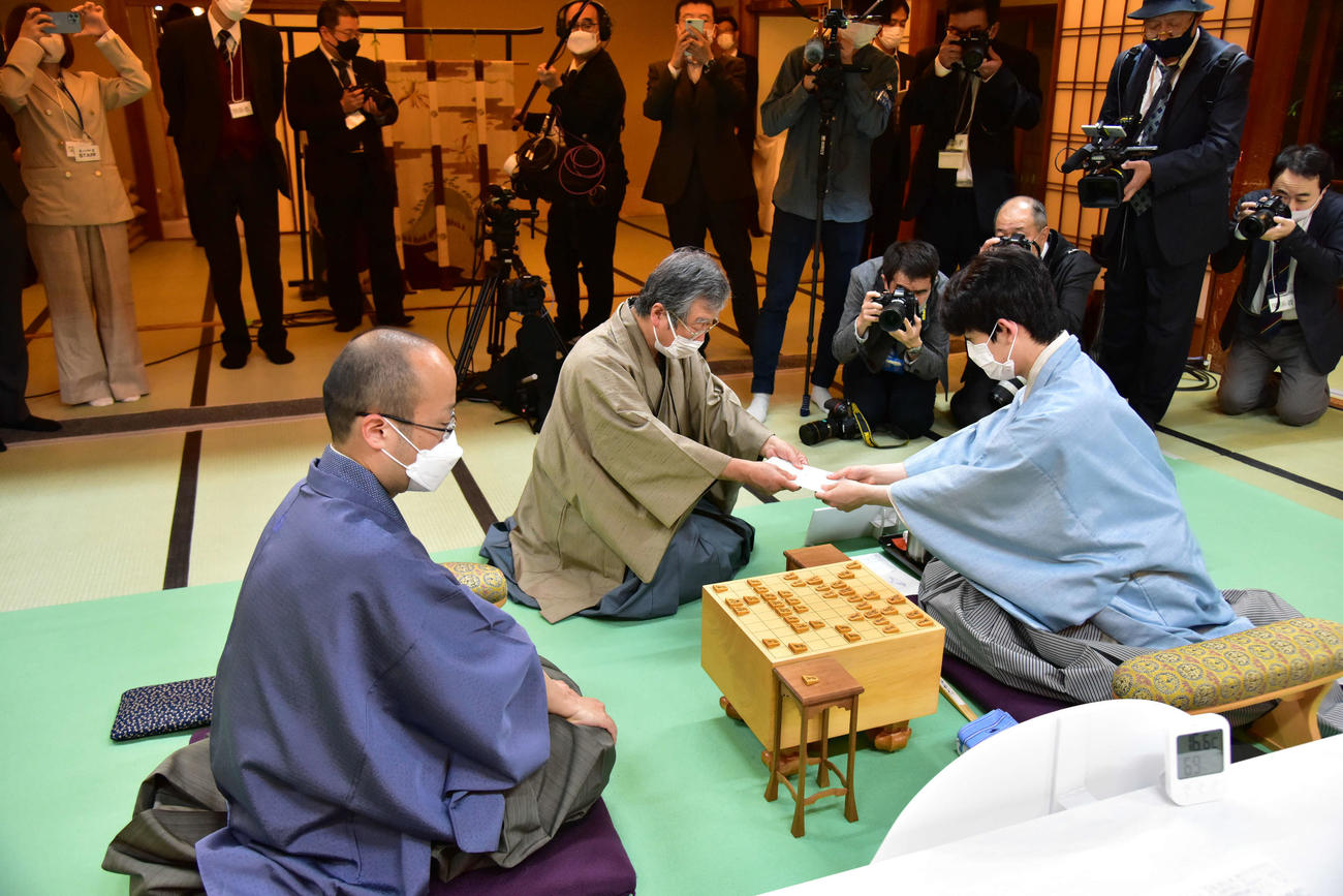 藤井聡太竜王（右）は封じ手の入った封筒を立会人の青野照市九段（中央）に手渡す（左は渡辺明名人、日本将棋連盟提供）