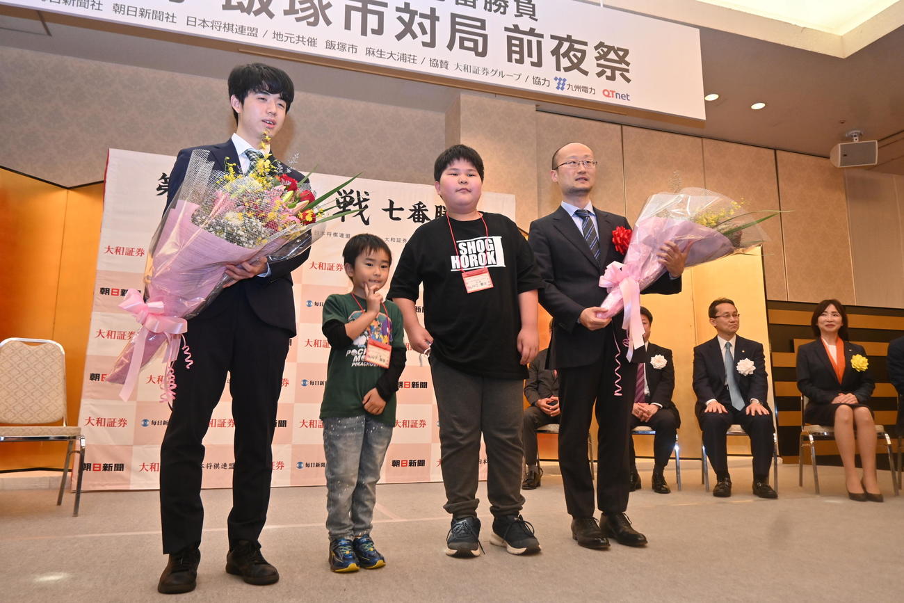 福岡県飯塚市内で開催された前夜祭で地元の小学生から花束を贈呈される藤井聡太竜王（左）渡辺明名人（撮影・松浦隆司）