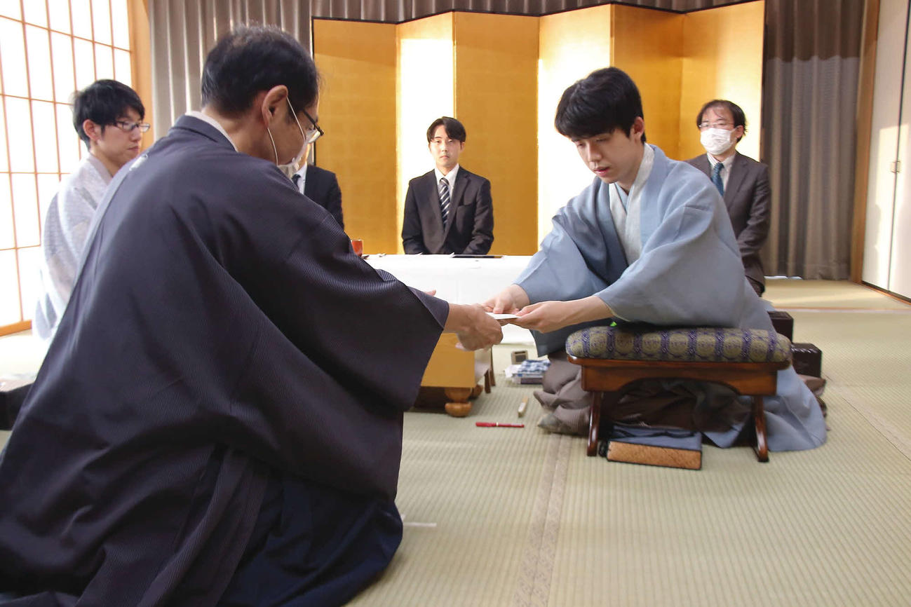 立会人の木村一基九段（中央）に封じ手の入った封筒を手渡す藤井聡太王位（右）。左は佐々木大地七段（日本将棋連盟提供）