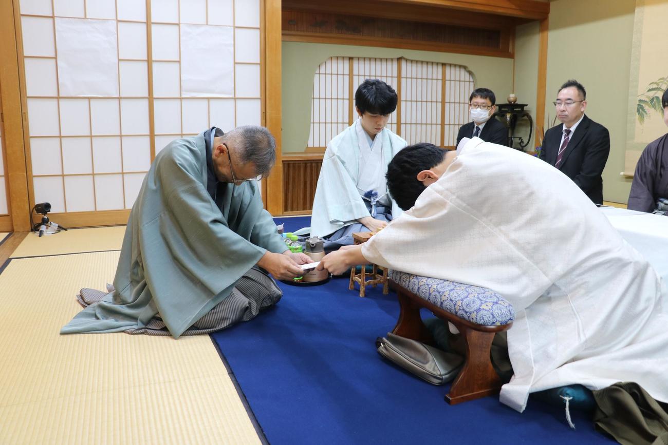 封じ手の封筒を立会人の中村修九段（左）に手渡す佐々木大地七段。奥は藤井聡太王位（日本将棋連盟提供）