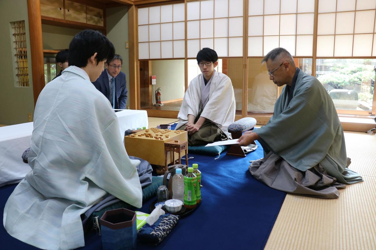 王位戦7番勝負第5局2日目、立会人の中村修九段が藤井聡太王位（手前）、佐々木大地七段に封じ手を見せる（日本将棋連盟提供）
