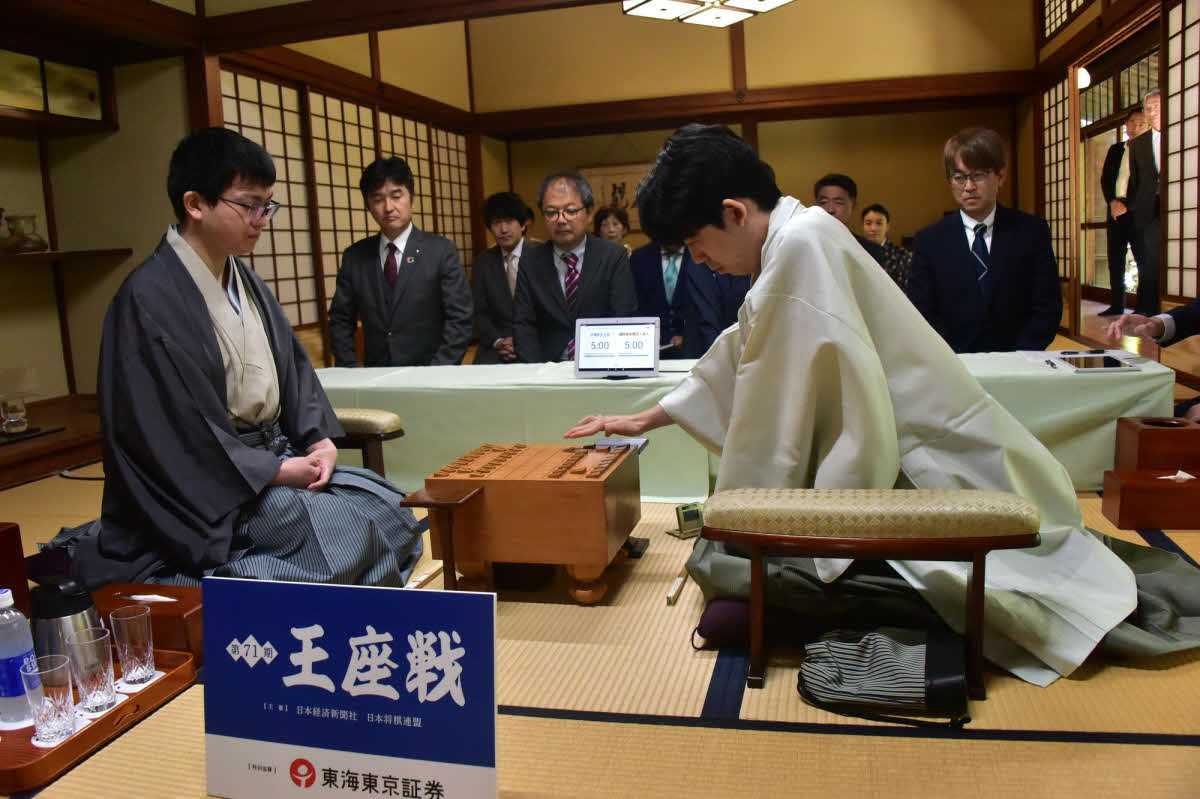 永瀬拓矢王座（左）に藤井聡太竜王（右）が初めて挑戦する王座戦第1局は藤井の先手でスタート（右後方は羽生善治会長、日本将棋連盟提供）